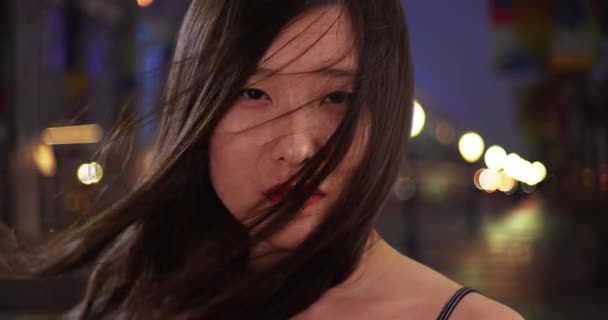 都市部の環境で風に吹いて髪を持つ素敵な千年紀の女性の閉じます 夜のシーンでカメラを見てアジアの女性の肖像画 — ストック動画