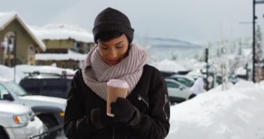 Güzel, genç bir kadın, elinde kahve ile karda, akıllı telefon kullanıyor. Kış günü cep telefonu kullanan neşeli gülümseyen siyah kadın. 4k