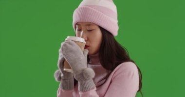 Kış mevsimi için giyinmiş, soğuktan donmuş Asyalı kadın yeşil ekrana çıkmak için sıcak tutuyor. Sıcak yeşil ekran stüdyosunu sıcak tutmak için sonbahar kıyafetleri ve sıcak bir içecek giyen hoş bir genç kadın. 4k