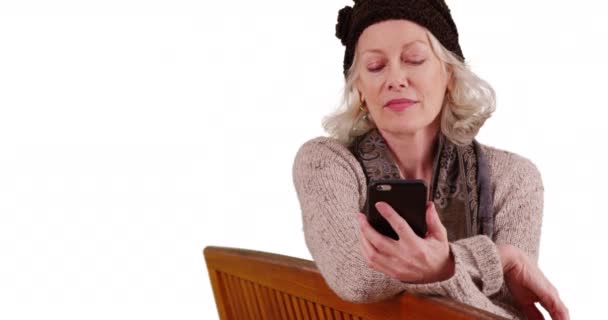 年长的女人坐在长椅上看着手机 在工作室里与复制空间混淆不清 迷人的老妇人用智能手机发短信与白底的回复相混淆 — 图库视频影像
