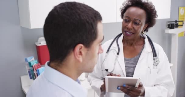 若いラテン系の患者と健康上の問題を議論するアフリカ系アメリカ人女性医師の肖像画 患者の健康履歴を見るために電子ノートパッドを使用して黒のシニア医療医師 — ストック動画