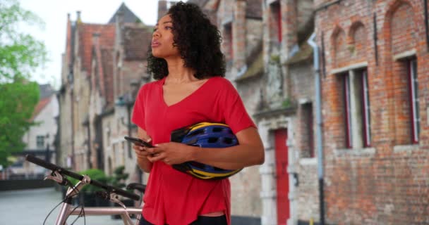 アフリカ系アメリカ人女性が携帯電話の地図アプリをチェックしている 自転車に乗って携帯電話を使用してベルギーで休暇中の黒人女性観光客 — ストック動画