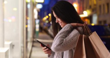 Alışveriş torbaları taşıyan güzel bir kadın geceleri şehir caddesinde selfie çekiyor. Akşamları akıllı telefonlu bir kadın fotoğraf çekiyor. 4k