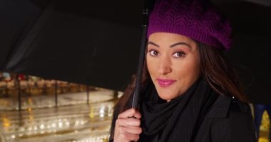 Yağmurlu bir gecede şehirde güzel bir Latin kadının portresi. Yağmurdan korunan rahat şapkalı ve eşarplı gülümseyen bir kadın. 4k
