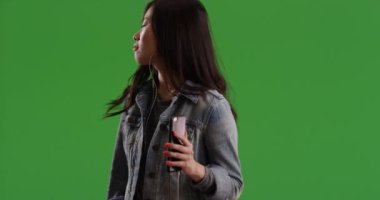 Neşeli Asyalı kadın yeşil ekranda müzik dinlemek için kulaklık takıyor ve akıllı telefon kullanıyor. Cep telefonlu mutlu genç bir kadın yeşil krom anahtar kompozitinde müziğin tadını çıkarıyor. 4k