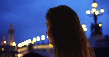 Pont Alexandre Köprüsü 'nün kenarında, Seine nehrine bakan güzel genç bir kadının yan görüntüsü. Kadın turistlerin gece gezintisi ve Paris 'i keşfetme silueti. 4k