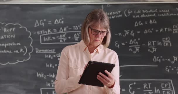 独立したシニア女性教師は 黒板の前でタブレット端末に取り組んでいます 黒板の前の教室でポータブル技術を楽しむ熟女 — ストック動画
