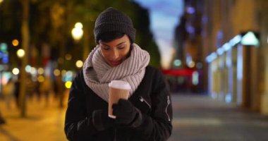 Akıllı telefon kullanarak bir fincan sıcak kahve içen güzel bir Afro-Amerikalı kadın. Geceleri dışarıda gülümseyen siyah bir kadın, telefon mesajlarına bakıyor. 4k