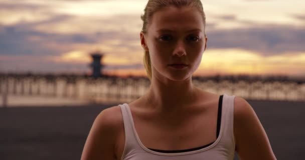日落时站在海滩上的女人 背景是码头 美丽的金发千禧年的年轻女子晚上在岸上摆姿势 — 图库视频影像