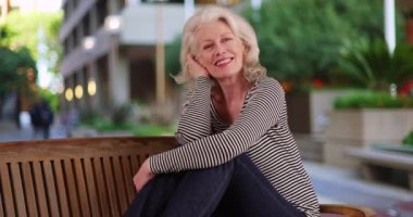Dışarıda bankta uzanmış kameraya gülümseyen sevimli son sınıf kadınının portresi. Kolları dizlerine dolanmış, kameraya gülen çekici yaşlı kadın. 4k