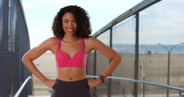 Güzel Afro-Amerikalı kadın, kumsalda güçlü, kaslı kollarıyla kameraya gülüyor. Sahilde elleri kalçalarında poz veren, spor kıyafetleri içinde güçlü bir kadın portresi. 4k