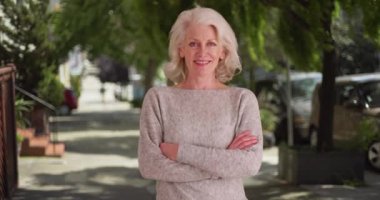 50 'li yaşlardaki neşeli olgun kadın portresi sessiz caddenin dışında kameraya gülümsüyor. Güzel yaşlı kadın kollarını açık havada kavuşturup gündüz vakti kameraya bakıyor. 4k