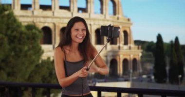 Roma Kolezyumu 'nun önünde selfie çubuğuyla gezen tatlı esmer kız. Roma 'daki neşeli turist kadın, arka planında ünlü bir simge olan fotoğraflar için poz veriyor. 4k