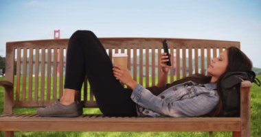 Beyaz kadın Golden Gate Köprüsü 'nün yanındaki park bankında cep telefonuyla mesajlaşıyor. Kot ceketli hippi kadın başının altında sırt çantasıyla bankta uzanıyor ve kahve tutuyor. 4k