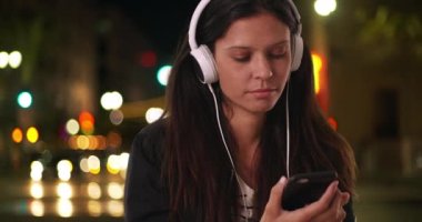 Cool Millennial 'lı bir kadın gece sokakta müzik dinlerken akıllı telefondan mesaj atıyor. Akşam şehir merkezinde cep telefonuyla mesajlaşan hippi kızı kapat. 4k