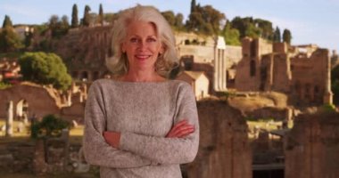 Roma harabelerinin önünde kolları çapraz duran mutlu son sınıf kadın turist. Gülümseyen olgun beyaz kadın İtalya 'da tatildeyken kameraya bakıyor. Roma 'da tatil. 4k