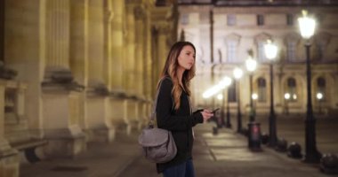 Paris caddesinde güzel beyaz bir kadın gece etrafa bakıyor. Genç bir kadın yol tarifi için akıllı telefondaki uygulamayı kullanıyor. 4k