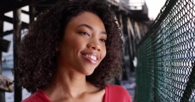 Şirin Afrikalı kadın okyanus rıhtımında gülümseyen ve kızaran özel birini düşünüyor. Genç bir kadın yeni sevgilisini düşünüyor. Mutlu hissediyor. Arka planda dalgalar çarpıyor. 4k