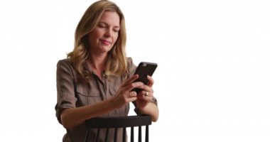 Orta yaşlı beyaz bir kadın beyaz arka planda akıllı telefondan kopya uzay ile mesajlaşıyor. Stüdyoda fotokopi odası olan sandalyede oturan bir kadın cep telefonu kullanıyor. 4k