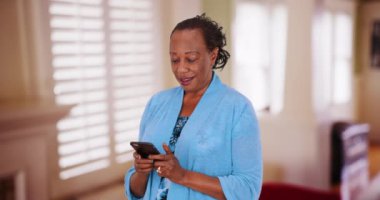 Siyah, yaşlı bir kadın mesaj atmak için akıllı telefonunu kullanıyor. Yaşlı bir Afrikalı Amerikalı kadın cep telefonunu evde kullanıyor. 4k