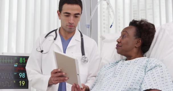 アフリカの女性患者の検査結果を示すために電子ノートブックタブレットを使用して魅力的な若いラテン系医師 シニア黒女性嘘で病院見でポータブルタブレットコンピュータ — ストック動画