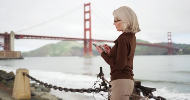 Profil Wanita Tua Berdiri Dengan Sepeda Browsing Ponsel Dekat Jembatan — Stok Video
