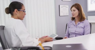 Genç bayan doktor tablet kullanıyor ve yaşlı meslektaşımla sağlık sigortası hakkında konuşuyor. İki kadın sağlık uzmanı, doktorların ofisinde farklı sağlık planları hakkında sohbet ediyorlar.