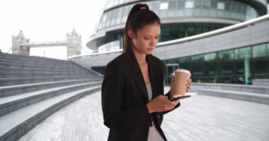 Milenyum işkadını çay içiyor ve cep telefonuyla mesajlaşıyor. Genç, profesyonel bir kadın, iş yeri parkında kahve fincanı içmek için mesaj atıyor ve gazete tutuyor. 4k