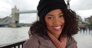 Tower Bridge 'in önünde ceketli ve bereli kameraya gülümseyen neşeli Afrikalı Amerikalı kadının yakın çekimi. Londra 'da tatilde olan Black Millennial turisti kış boyunca Thames nehrinin yanında duruyor. 4k