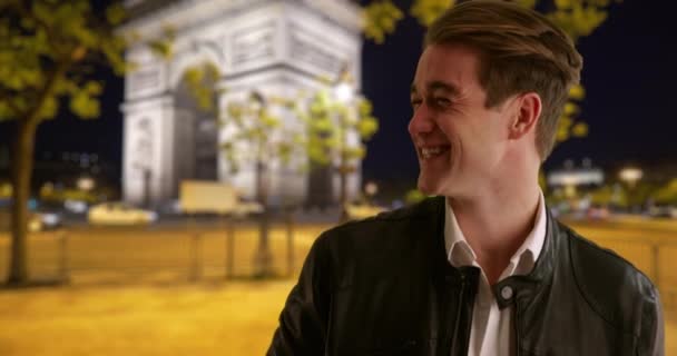 一个英俊的年轻人晚上在巴黎街上笑着 穿着皮夹克的又酷又时髦的家伙在镜头前眨眼 — 图库视频影像
