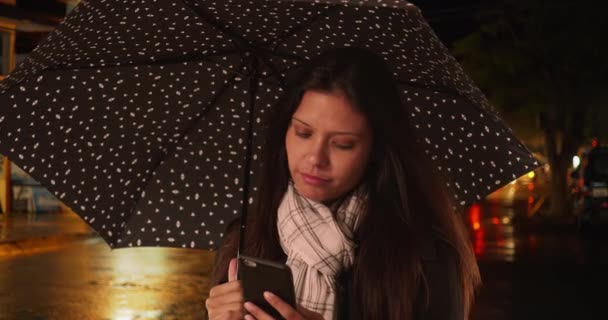 路上の傘の下のスマートフォンでテキストメッセージを笑顔で読む100万人の女性 携帯電話を使用して夜の暴風雨の間 都市部の女性 — ストック動画