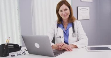 Ofis masasında oturmuş kameraya gülümseyen güzel bir bayan doktorun portresi. Kameraya bakan profesyonel tıp hekimine yakın çekim