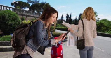 Arkadaşı İtalya 'daki bir haritaya bakarken çekici beyaz kadın cep telefonuyla selfie çekiyor. Arkadaşı kendi fotoğrafını çekerken sarışın kadın turist seyahat haritasına bakıyor. 4k