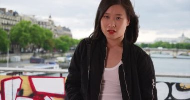 Siyah bombardıman ceketli Çinli kadın Seine nehrinin üzerinde, rüzgarda savrulan saçlarıyla köprüde. Asyalı milenyum turisti Paris 'te cepleriyle kaldırımda bekliyor. 4k