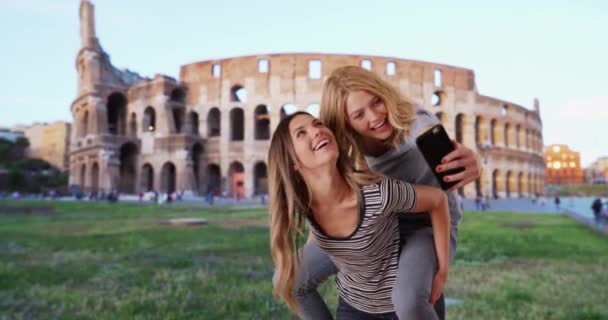 彼女の友人の背中にピギーバックに乗って魅力的なブロンドの白人女性は ローマコロッセオ近くの自撮りを取ります ローマのかなりブルネットは自分自身を取るために友人のピギーバックに乗るを与えます — ストック動画