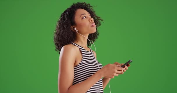 身穿黑白相间条纹衬衫的女人 一边听音乐 一边用带有绿屏背景的智能手机 非裔美国千禧年女性使用短信应用和佩戴绿色耳塞 — 图库视频影像