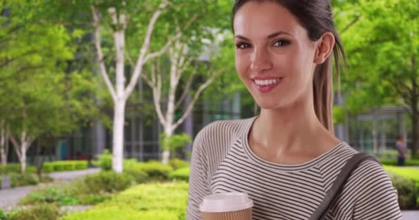 建物の中庭でコーヒーを保持幸せな若い白人女性の肖像画 現代的な建物の外のカメラで笑みを浮かべてコーヒーカップと彼女の20代の若いブルネット — ストック動画