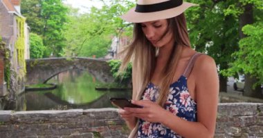Belçika, Bruges 'e seyahat ederken, Millennial Girl in Romper ve Fötr şapkalı mesaj atıyor. 20 'li yaşlarda çekici bir kız, Bruges' deki köprüde mesaj göndermek için akıllı telefon kullanıyor. 4k