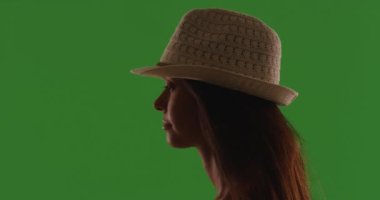 Yeşil perdenin önünde fötr şapka takan çekici genç bir kadının silueti. Profil görüntüsü: Şapkalı ve saçı yeşil ekranda uçuşan güzel bir bayan. 4k
