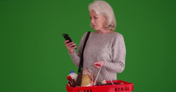 緑の画面上の携帯電話でシニア白人女性のショッピングやメッセージングの肖像画 緑の画面上の電話でテキストメッセージを食料品でいっぱいのバスケットを保持する女性は キーまたは構成される — ストック動画