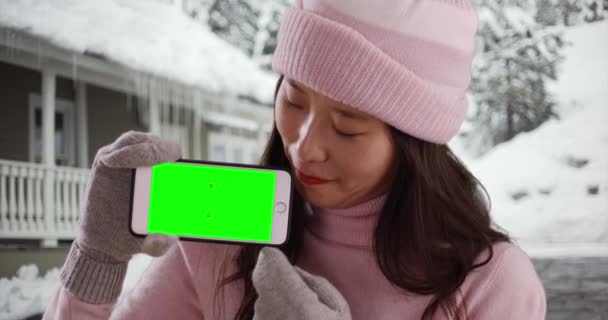 在下雪天 穿着冬衣的中国女人拿着装有绿色屏风的智能手机微笑 快乐的一千年与手套和毛衣举起手机显示屏幕和看它 — 图库视频影像