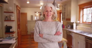 50 'li yaşlarda beyaz bir kadın. Büyüleyici mutfağın içinde kameraya gülümseyen kollarını kavuşturmuş poz veriyor. Evde kameraya gülümseyen gri kazaklı neşeli yaşlı kadın. 4k