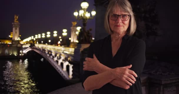 在法国巴黎 一位严肃的老年妇女抱着胳膊 在夜里严肃地看着外面的摄像机 巴黎亚历山德拉三世桥的一位漂亮而成熟的女人 她有着严肃的眼神 交叉着双臂 — 图库视频影像