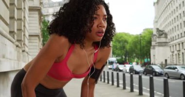 Afro-Amerikalı kadın koşucu soluklanıyor ve fitness izleyicisini kontrol edip ekrandan çıkıyor. Siyah kadın koşucu, kulaklıkları takıp kaldırımda dinleniyor sonra da koşuyor. 4k