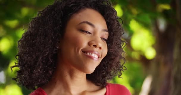 可爱的非洲女性在想一个特别的微笑的人 年轻的女人站在树下 绿叶鲜亮 感到温暖和快乐 — 图库视频影像