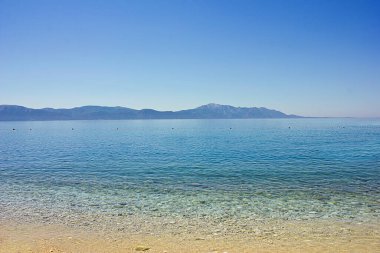 Altından plajı ve ufukta dağları olan turkuaz bir deniz.