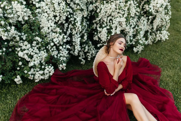 Piękna Dziewczyna Czerwonej Sukience Siedzi Pobliżu Krzaka Białymi Kwiatami — Zdjęcie stockowe