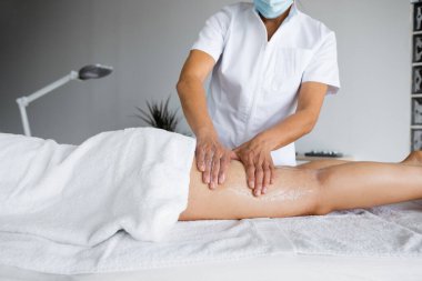 Crop unrecognizable beautician wearing protective face mask, rubbing scrub gel into leg skin of woman during anti cellulite massage session on table in parlor