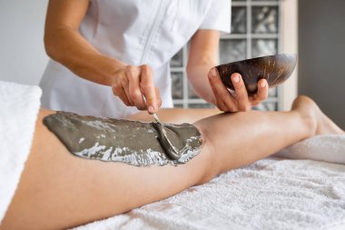 Crop beautician applying black clay on leg of unrecognizable barefoot woman on table in spa salon. close up, detail of woman leg with treatment on it. Horizontal