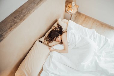Top view of Young brunette woman sleeping peacefully on cozy bed in morning hugging soft beige pillow, fresh soft bedding white linen and mattress in bedroom. Good night sleep concept. Horizontal